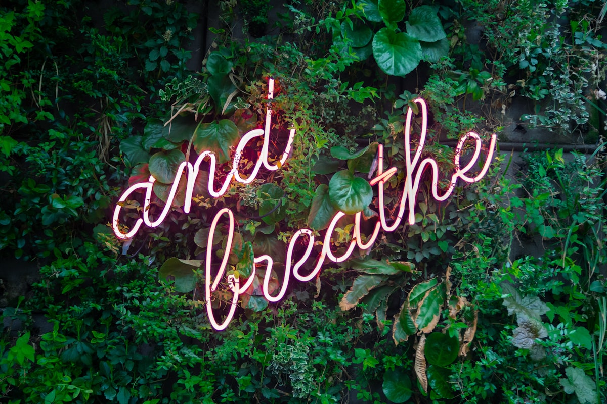 Neon sign reading 'and breathe' surrounded by lush green plants